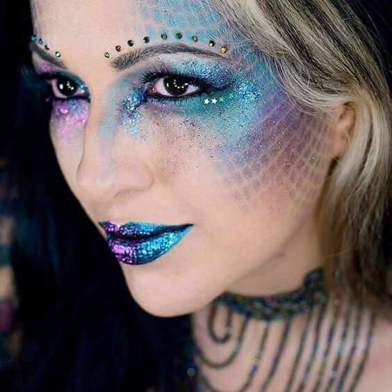 Mulher loira com maquiagem de sereia, glitter colorido ao redor dos olhos, boca colorida e pedrinhas acima das sobrancelhas