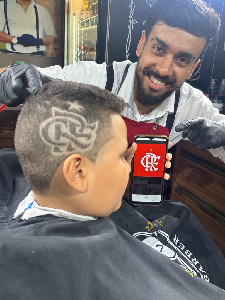 KF em um corte freestyle reproduzindo no cabelo do cliente o símbolo do flamengo, ao lado, ele segurando um celular com o símbolo do clube aberto 