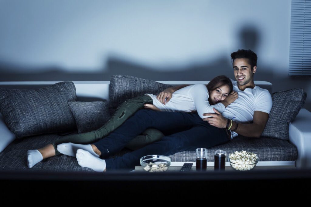 Casal vendo filmes juntos, os dois sentados no sofá, o homem de camisa branca encostado no braço do sofá com uma almofada, a mulher de camiseta manga longa branca deitada em seu colo