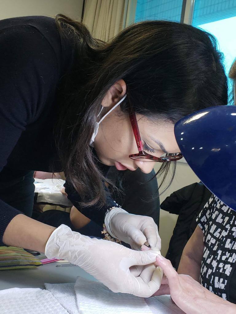 Mulher de avental preto, luva branca, cabelo preto, com óculos vermelho cortando uma unha de uma mão branca