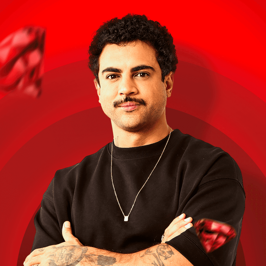 Homem branco de cabelo preto e bigode com os braços cruzados, corpo inclinado para a esquerda, ao fundo um diamante vermelho e o fundo vermelho, outro diamante vermelho na manga de sua camiseta